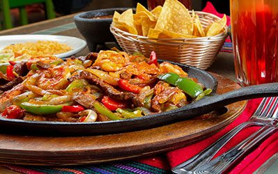 Combination chicken, shrimp and beef fajitas from a gourmet Mexican restaurant.  Food is presented in a rustic cast iron fajita skillet with a wooden base and also includes onions, spices and bell pepper.  Main dish is displayed on a table with chips, salsa, guacamole and Iced tea with lemon.