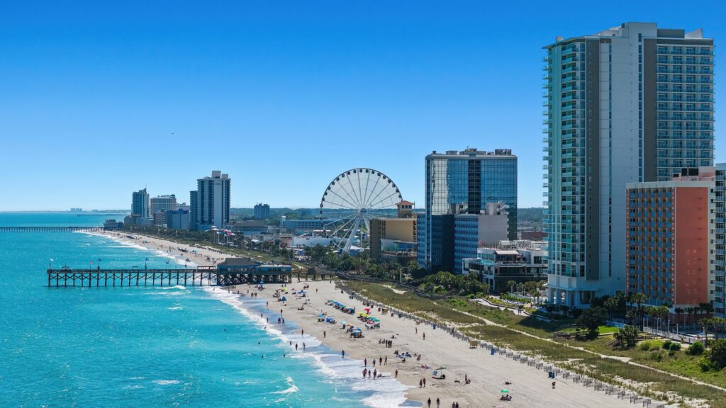Aerial view of the Myrtle Beach coastline on a clear day, showing turquoise ocean waves, a wide sandy beach dotted with beachgoers and colorful umbrellas, a wooden fishing pier extending into the water, the SkyWheel Ferris wheel, and a row of high-rise hotels and condominiums lining the shore.